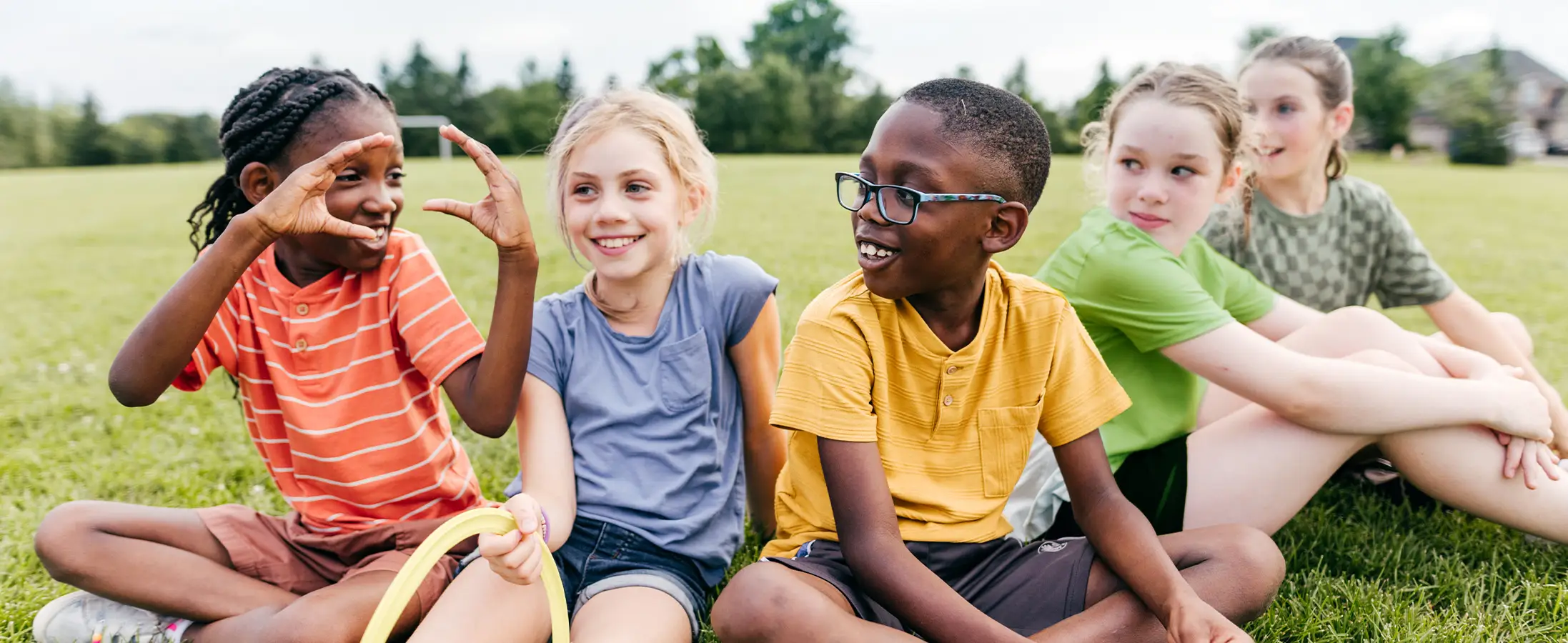 Five elementary age students are sitting outside talking with each other and smiling.
