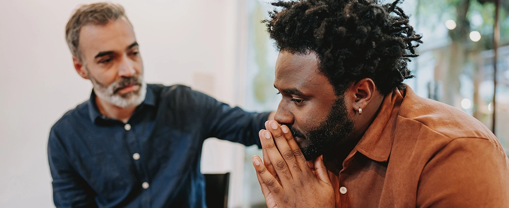 Man in a navy button down shirt resting his hand on another man's shoulder.