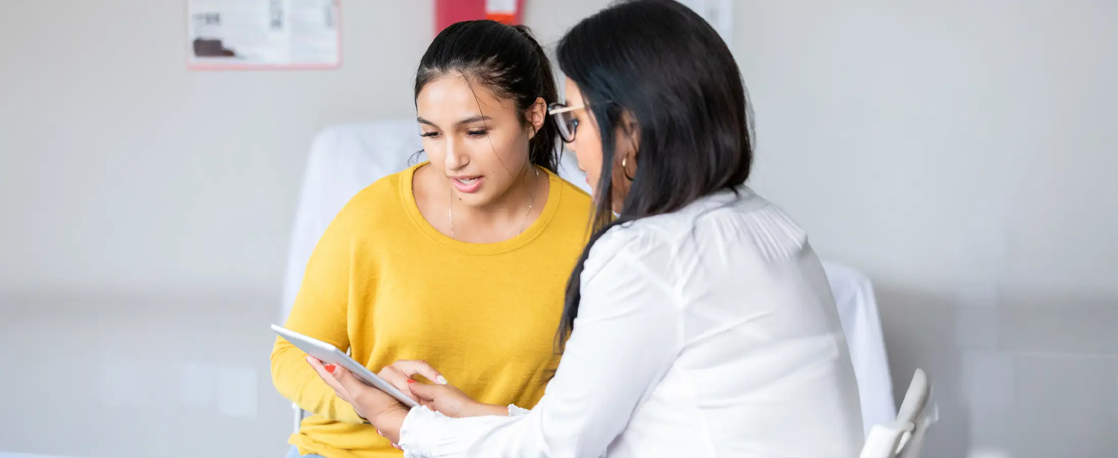 A woman in a white long sleeve shirt and glasses is showing another woman in a yellow long sleeve shirt her ipad screen.