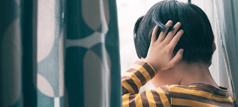 A young boy/child looks out a window covering his ears to represent childhood trauma