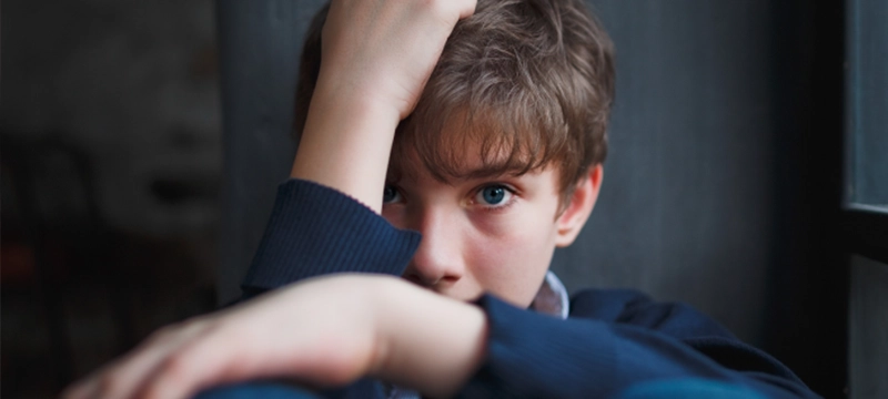 A young boy hides his face while crouching by a window, representing childhood trauma