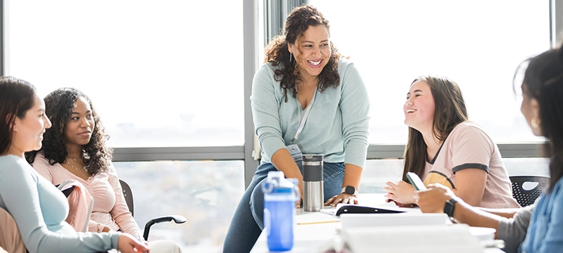 A teacher smiles while a group of female students sit around her smiling in a lively discussion, emphasizing positive mental health and supporting students as they return back to school