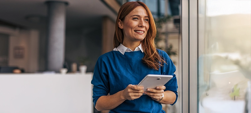 A redheaded woman smiles looking out a window in an office setting while holding a tablet, representing how psychologists can save time with digital tools like the RIAS-2 DRF