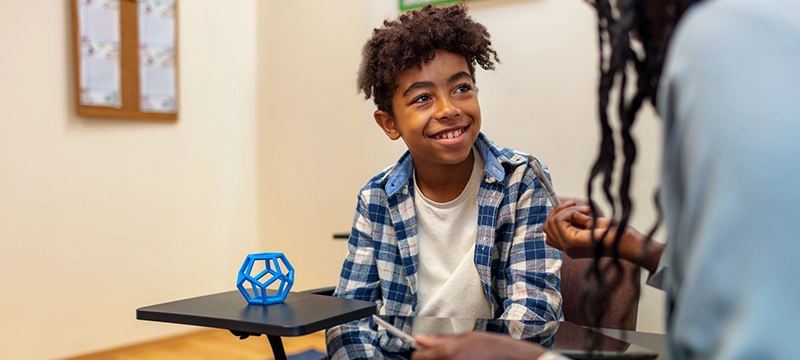 A young boy with curly hair smiles at his teacher or school psychologist who gestures in an explanatory manner. The boy sits with a fidget toy on his desk, symbolizing ADHD.